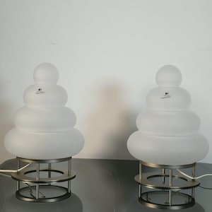 Può includere: Due lampade da tavolo con globi in vetro bianco satinato impilati in una forma arrotondata e a gradini. Ogni lampada poggia su una base circolare in metallo spazzolato sostenuta da quattro aste metalliche verticali. Etichetta nera visibile.