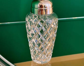 May include: A clear glass cocktail shaker with a diamond-cut pattern. The shaker has a silver-colored lid and top. The glass is reflecting light. The background is a solid green color.