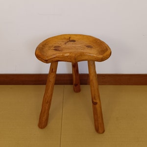 Può includere: Uno sgabello in legno rustico con tre gambe e una seduta arrotondata a forma di sella. Lo sgabello è in legno naturale con una finitura marrone chiaro.