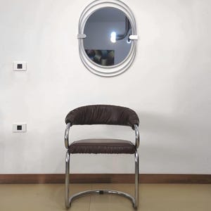May include: A brown leather chair with a chrome frame. The chair has a curved back and a rounded seat. The chair is in front of a white wall with a silver oval mirror with multiple concentric circles.