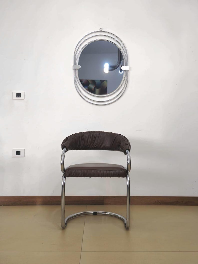 May include: A brown leather chair with a chrome frame. The chair has a curved back and a rounded seat. The chair is in front of a white wall with a silver oval mirror with multiple concentric circles.