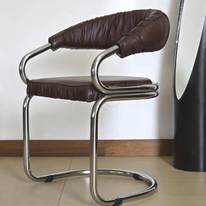 May include: A brown leather chair with a chrome frame. The chair has a curved back and a padded seat. The chair is on a tiled floor.