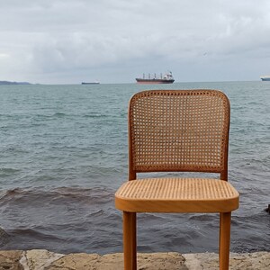 May include: A wooden chair with a woven cane seat and back, sitting on a stone path overlooking a body of water. The chair is facing the water, with a large ship in the distance.