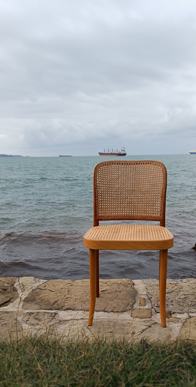 May include: A wooden chair with a woven cane seat and back, sitting on a stone path overlooking a body of water. The chair is facing the water, with a large ship in the distance.