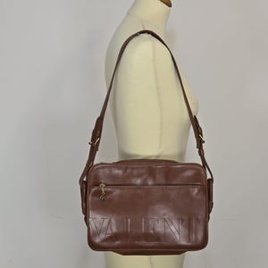 Pode incluir: Bolsa transversal de couro marrom com a palavra "VALENTINO" em relevo na frente. A bolsa tem uma longa al&ccedil;a ajust&aacute;vel e ferragens douradas. A bolsa &eacute; exibida em um manequim.