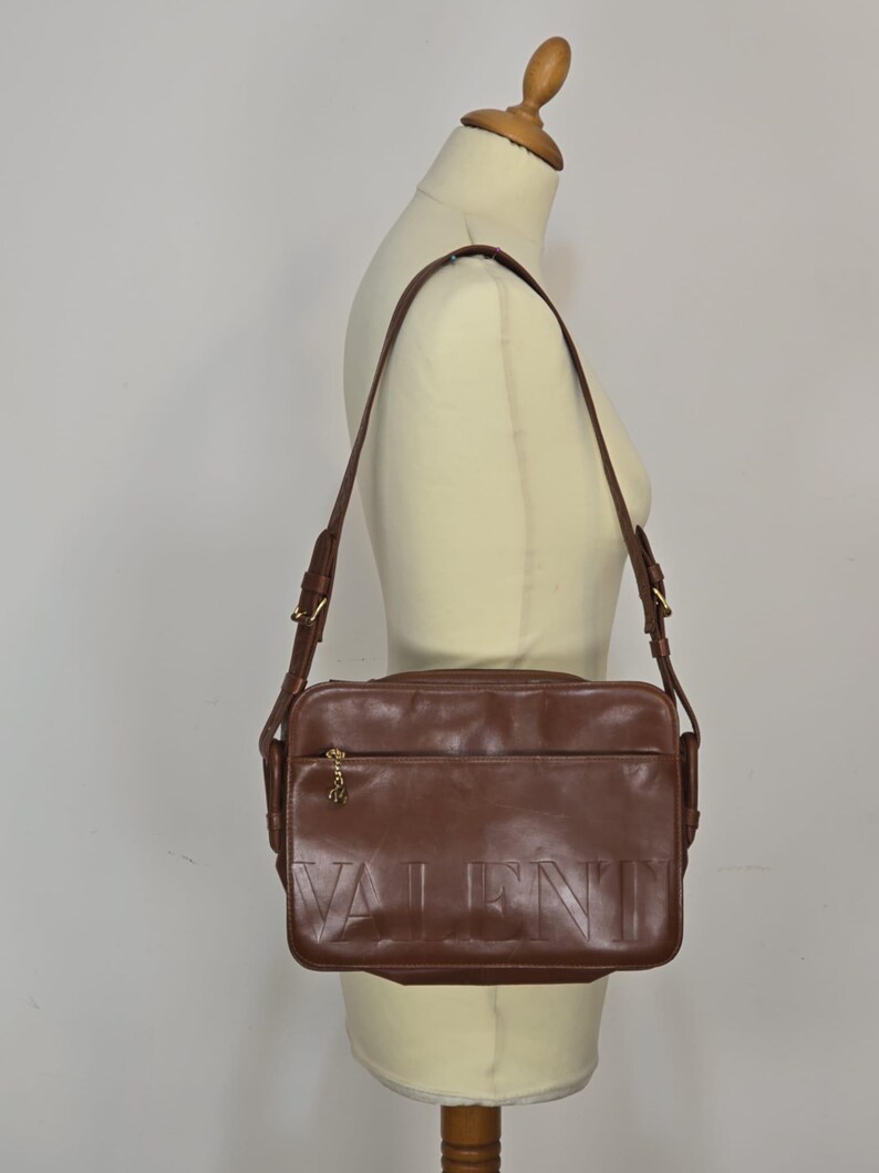 Pode incluir: Bolsa transversal de couro marrom com a palavra "VALENTINO" em relevo na frente. A bolsa tem uma longa al&ccedil;a ajust&aacute;vel e ferragens douradas. A bolsa &eacute; exibida em um manequim.