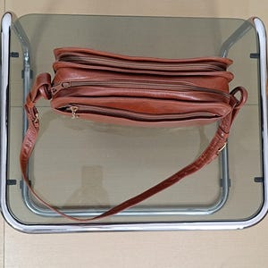 Pode incluir: Uma bolsa transversal de couro marrom com v&aacute;rios compartimentos com z&iacute;per. A bolsa tem uma al&ccedil;a longa e ajust&aacute;vel e ferragens douradas. O couro parece ligeiramente desgastado, dando-lhe um aspecto vintage. A bolsa est&aacute; sobre uma superf&iacute;cie de vidro.