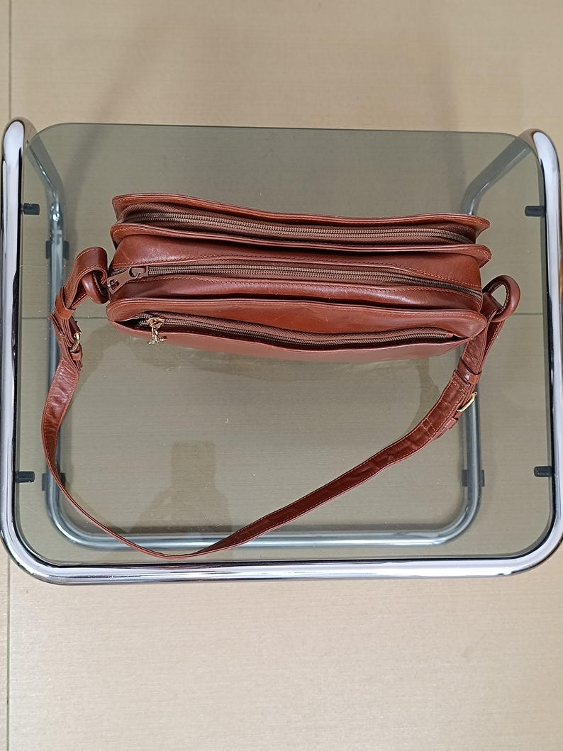 Pode incluir: Uma bolsa transversal de couro marrom com v&aacute;rios compartimentos com z&iacute;per. A bolsa tem uma al&ccedil;a longa e ajust&aacute;vel e ferragens douradas. O couro parece ligeiramente desgastado, dando-lhe um aspecto vintage. A bolsa est&aacute; sobre uma superf&iacute;cie de vidro.