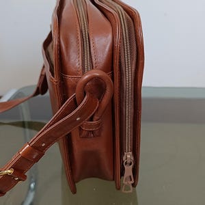Pode incluir: Uma bolsa transversal de couro marrom com v&aacute;rios compartimentos com z&iacute;per. A bolsa possui uma al&ccedil;a de ombro, uma al&ccedil;a e uma fivela dourada. A bolsa &eacute; feita de um material de couro liso e brilhante.