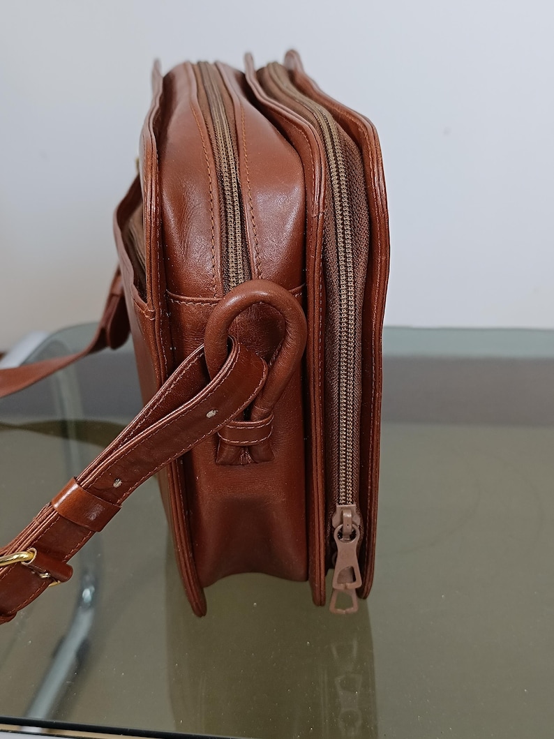 Pode incluir: Uma bolsa transversal de couro marrom com v&aacute;rios compartimentos com z&iacute;per. A bolsa possui uma al&ccedil;a de ombro, uma al&ccedil;a e uma fivela dourada. A bolsa &eacute; feita de um material de couro liso e brilhante.