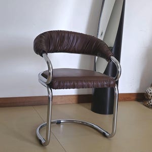May include: A brown leather chair with a chrome frame. The chair has a curved back and a padded seat. The chair is sitting on a tiled floor.
