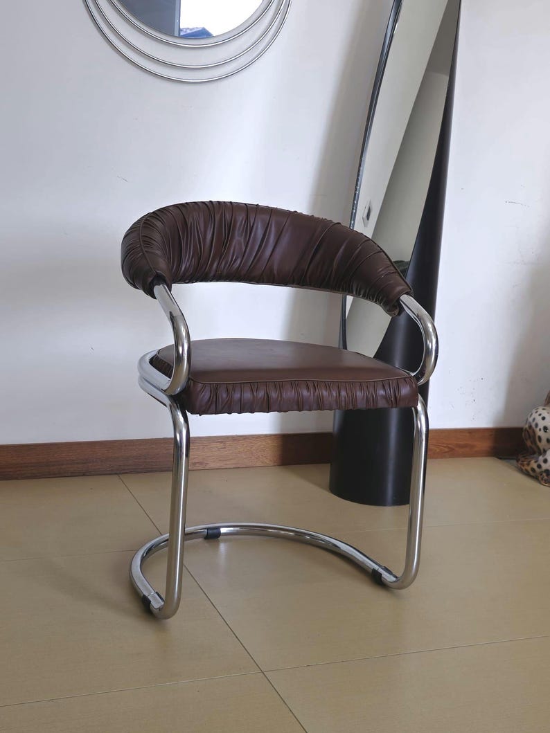 May include: A brown leather chair with a chrome frame. The chair has a curved back and a padded seat. The chair is sitting on a tiled floor.