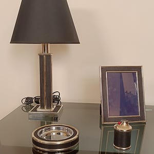 Peut inclure: Une lampe noire et argentée avec un abat-jour noir est posée sur une table en verre. Un cendrier, un cadre photo et un briquet assortis sont également présents. Les articles ont des accents argentés.