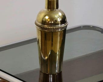 Puede incluir: Un coctelera de oro con una banda plateada alrededor del medio. La coctelera está sentada en una mesa de vidrio con un marco de cromo.