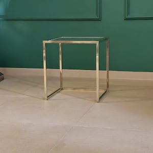 May include: A gold metal side table with a square glass top. The table is sitting on a beige tiled floor in front of a green wall.