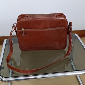 Pode incluir: Bolsa transversal de couro marrom com o nome da marca "VALENTINO" em relevo na frente. A bolsa tem uma al&ccedil;a longa e ajust&aacute;vel e v&aacute;rios compartimentos com z&iacute;per. Um pingente dourado pende do z&iacute;per.
