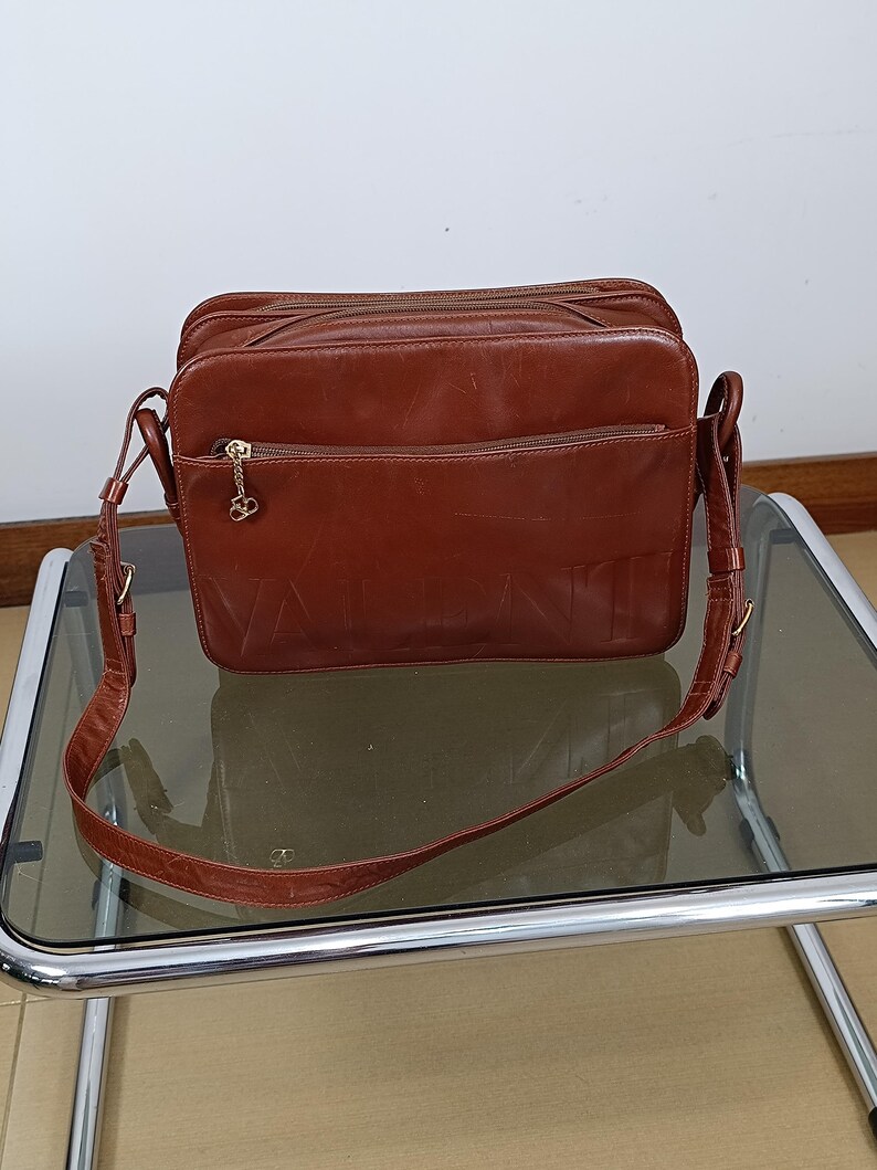 Pode incluir: Bolsa transversal de couro marrom com o nome da marca "VALENTINO" em relevo na frente. A bolsa tem uma al&ccedil;a longa e ajust&aacute;vel e v&aacute;rios compartimentos com z&iacute;per. Um pingente dourado pende do z&iacute;per.