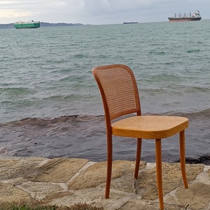 May include: A wooden chair with a woven cane seat and back, sitting on a stone path by the water's edge. The chair is facing the ocean, with a cloudy sky and two ships in the distance.