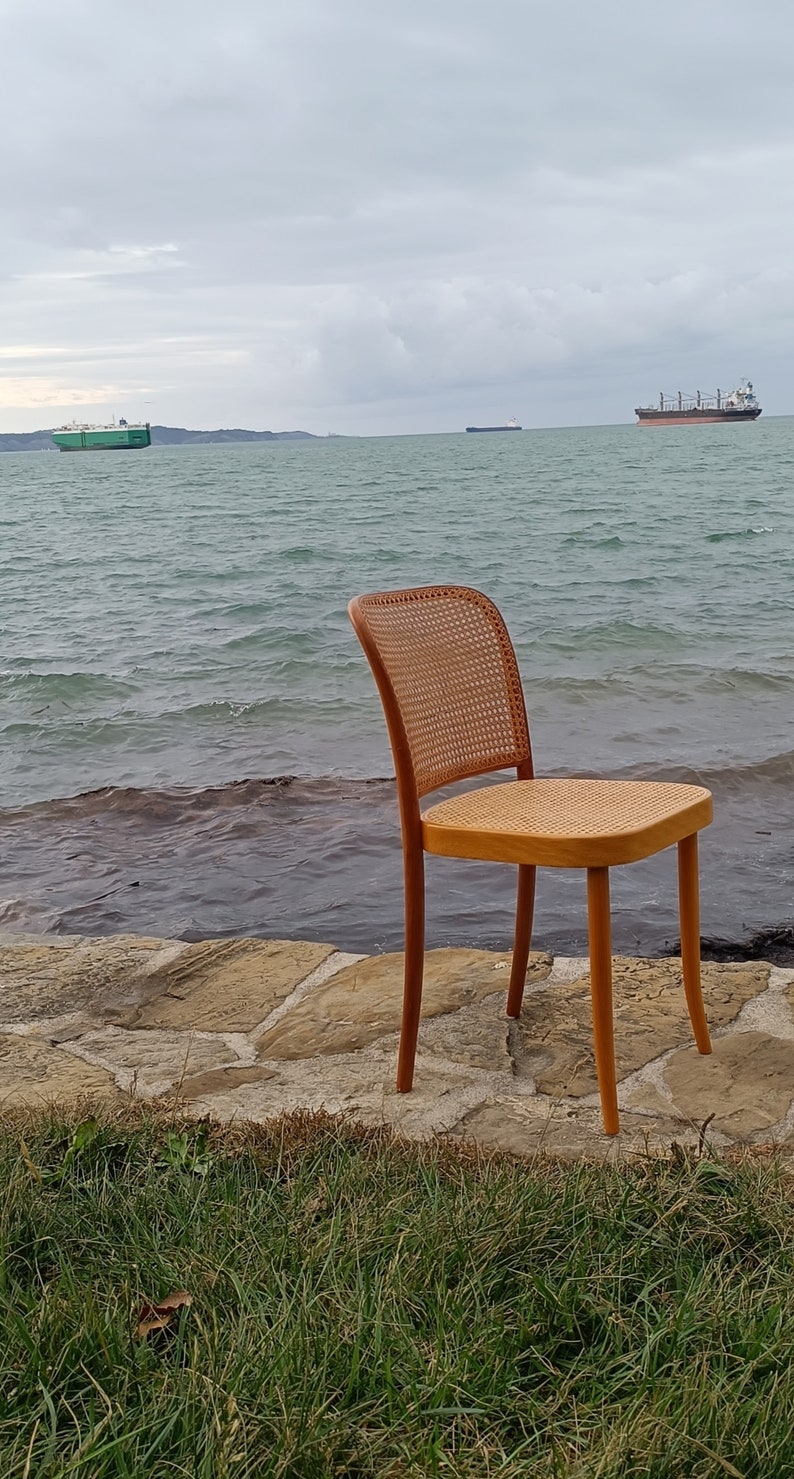 May include: A wooden chair with a woven cane seat and back, sitting on a stone path by the water's edge. The chair is facing the ocean, with a cloudy sky and two ships in the distance.