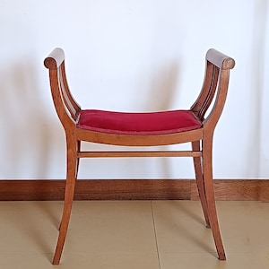 Op de afbeelding: Een houten kruk met een rood fluwelen kussen. De kruk heeft een gebogen rugleuning en vier poten.