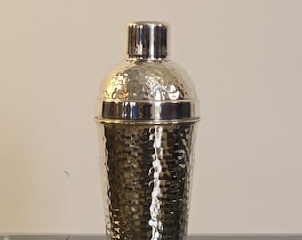 Puede incluir: Un coctelera de plata con acabado martillado. La coctelera tiene una parte superior redonda y un cuerpo cilíndrico.