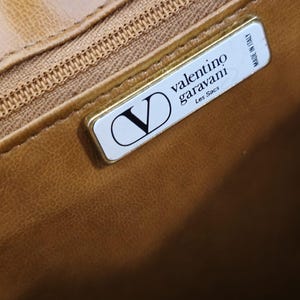 Pode incluir: Close-up do interior de uma mala de couro castanho com um fecho dourado e uma etiqueta retangular. A etiqueta apresenta o log&oacute;tipo Valentino Garavani, as palavras "Les Sacs" e "Made in Italy".
