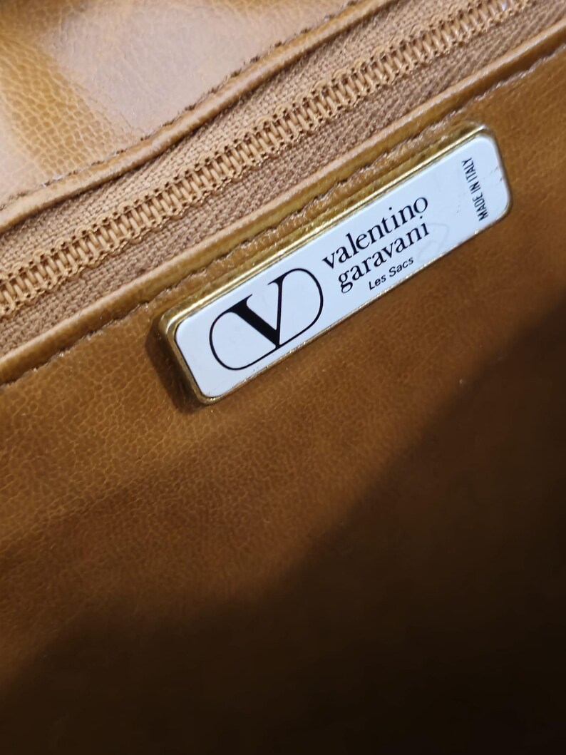 Pode incluir: Close-up do interior de uma mala de couro castanho com um fecho dourado e uma etiqueta retangular. A etiqueta apresenta o log&oacute;tipo Valentino Garavani, as palavras "Les Sacs" e "Made in Italy".