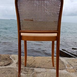 May include: A wooden chair with a woven cane back and seat. The chair is brown and has a simple design. It is sitting on a stone surface with grass in the foreground.