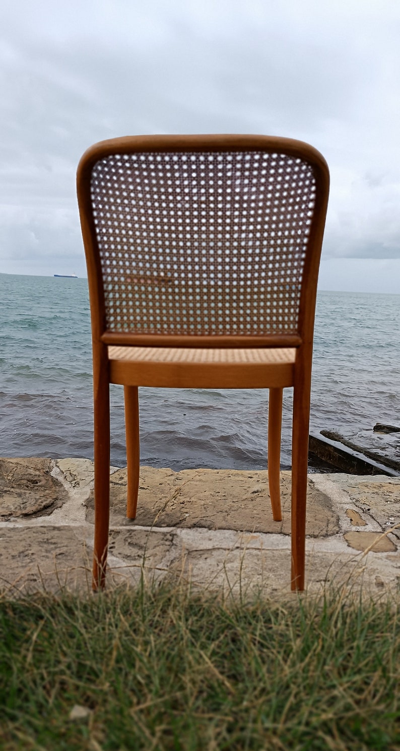 May include: A wooden chair with a woven cane back and seat. The chair is brown and has a simple design. It is sitting on a stone surface with grass in the foreground.