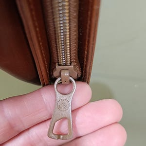 Pode incluir: Close-up de uma carteira de couro castanho com z&iacute;per. O puxador do z&iacute;per &eacute; de metal castanho claro com as letras "CC" dentro de um c&iacute;rculo. A carteira est&aacute; parcialmente aberta.