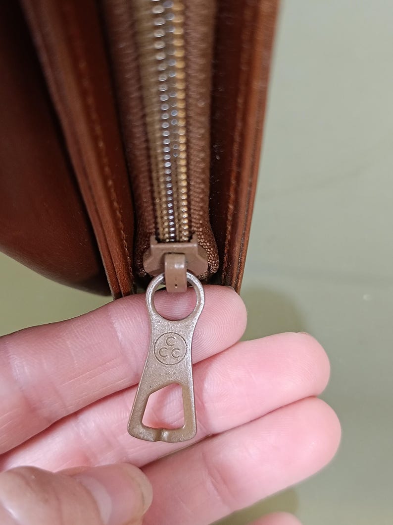 Pode incluir: Close-up de uma carteira de couro castanho com z&iacute;per. O puxador do z&iacute;per &eacute; de metal castanho claro com as letras "CC" dentro de um c&iacute;rculo. A carteira est&aacute; parcialmente aberta.