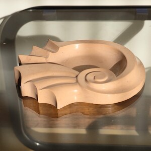 Può includere: Un oggetto decorativo a forma di conchiglia di colore pesca chiaro. La scultura ha un design a spirale con una superficie liscia e curva e un bordo strutturato. L'oggetto è appoggiato su una superficie di vetro riflettente.