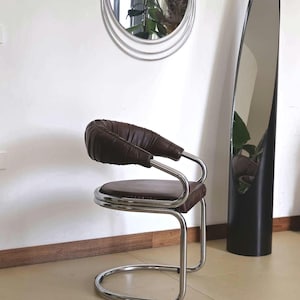 May include: A brown leather chair with a chrome frame. The chair has a curved back and a round seat. The chair is in front of a tall, oval mirror.