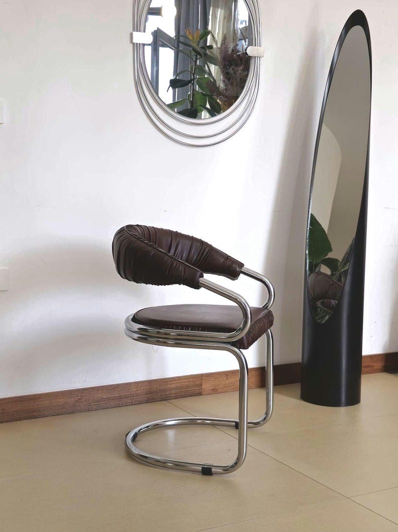 May include: A brown leather chair with a chrome frame. The chair has a curved back and a round seat. The chair is in front of a tall, oval mirror.