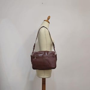 Pode incluir: Uma bolsa de ombro de couro marrom com uma al&ccedil;a longa ajust&aacute;vel. A bolsa retangular tem um z&iacute;per e um pequeno pingente decorativo. A bolsa &eacute; exibida em um manequim.