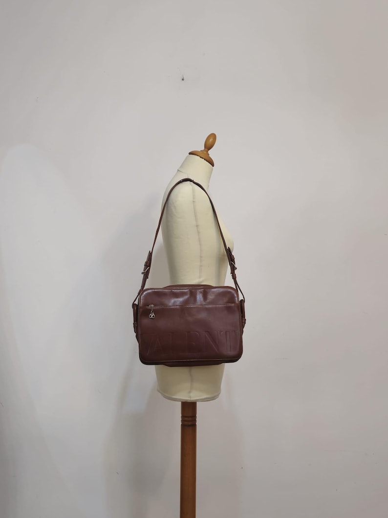 Pode incluir: Uma bolsa de ombro de couro marrom com uma al&ccedil;a longa ajust&aacute;vel. A bolsa retangular tem um z&iacute;per e um pequeno pingente decorativo. A bolsa &eacute; exibida em um manequim.