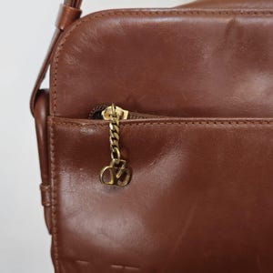 Pode incluir: Bolsa transversal de couro castanho com corrente dourada e um berloque decorativo. A bolsa possui um bolso frontal com z&iacute;per e uma al&ccedil;a de ombro. O couro tem uma textura lisa e um brilho sutil, sugerindo um estilo vintage.