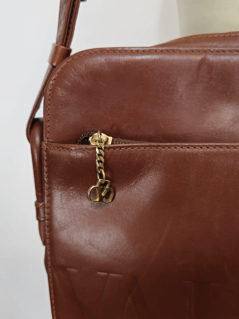 Pode incluir: Bolsa transversal de couro castanho com corrente dourada e um berloque decorativo. A bolsa possui um bolso frontal com z&iacute;per e uma al&ccedil;a de ombro. O couro tem uma textura lisa e um brilho sutil, sugerindo um estilo vintage.