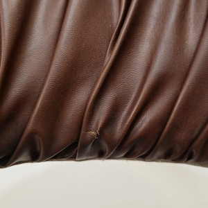 May include: Close-up of a brown leather fabric with a textured surface. The fabric is folded and has a slight wrinkle in the center.