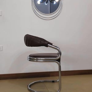 May include: A brown leather chair with a chrome frame. The chair has a curved back and a round seat. The chair is sitting on a light brown floor.