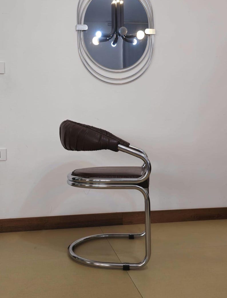 May include: A brown leather chair with a chrome frame. The chair has a curved back and a round seat. The chair is sitting on a light brown floor.