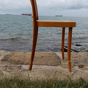 May include: A wooden chair with a woven seat and back sits on a rocky shore overlooking the ocean. The chair is made of light brown wood and has a simple design.
