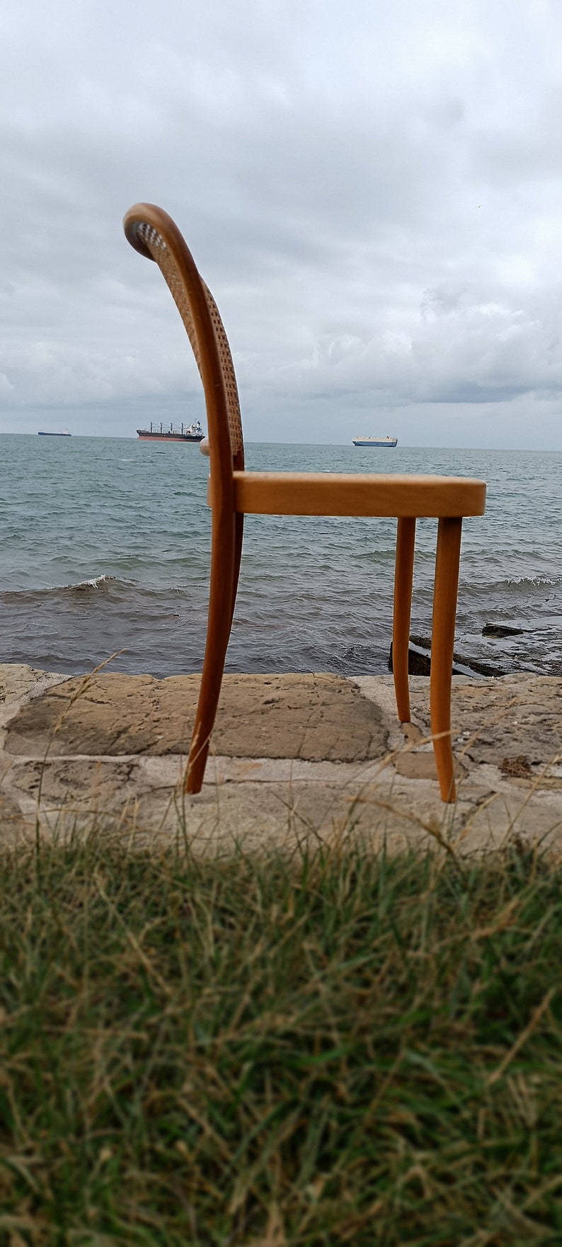 May include: A wooden chair with a woven seat and back sits on a rocky shore overlooking the ocean. The chair is made of light brown wood and has a simple design.