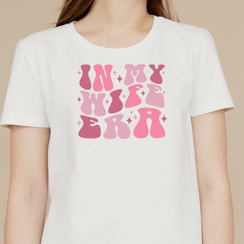 May include: White t-shirt with a pink and white graphic that says "In My Wife Era" in a retro font.