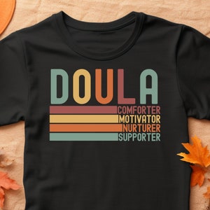 Op de afbeelding: Zwart T-shirt met het woord "Doula" in een retro-lettertype met kleurrijke strepen eronder. De tekst "comforter, motivator, nurturer, supporter" staat in wit onder de strepen.