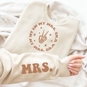 May include: A beige sweatshirt with the text "In My Mrs. Era" in a circle with a skeleton hand making a peace sign and stars. The text "Mrs." is also on the sleeve of the sweatshirt.