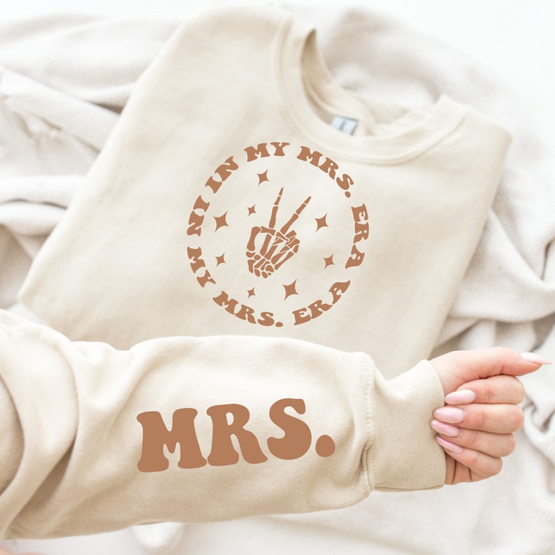 May include: A beige sweatshirt with the text "In My Mrs. Era" in a circle with a skeleton hand making a peace sign and stars. The text "Mrs." is also on the sleeve of the sweatshirt.