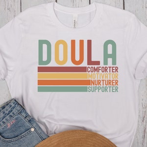 Op de afbeelding: Wit T-shirt met het woord "DOULA" in grote, kleurrijke letters. Onder het woord staan de woorden "COMFORTER", "MOTIVATOR", "NURTURER" en "SUPPORTER" in kleinere, kleurrijke letters.