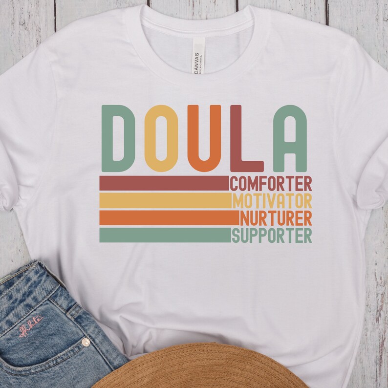 Op de afbeelding: Wit T-shirt met het woord "DOULA" in grote, kleurrijke letters. Onder het woord staan de woorden "COMFORTER", "MOTIVATOR", "NURTURER" en "SUPPORTER" in kleinere, kleurrijke letters.