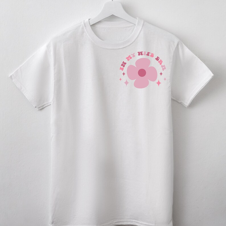 May include: White t-shirt with a pink flower graphic and the text "In My Wife Era" in a pink, rainbow-colored font.